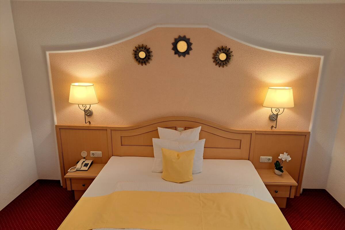 Une chambre d’hôtel avec un grand lit, deux lampes de chevet, un téléphone et une petite table sur laquelle se trouve un vase avec des fleurs.