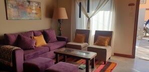 Ein Wohnzimmer mit einem violetten Sofa, zwei Sesseln, einem Glastisch, einer Lampe und einem Gemälde an der Wand.