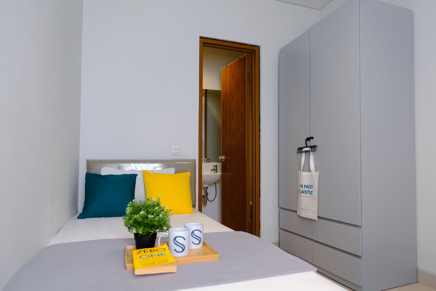A neatly arranged bedroom with a bed, a bedside table with a book, a potted plant, and two mugs.