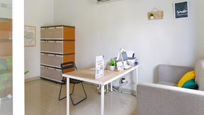 Habitación limpia y moderna con escritorio, computadora y zona de estar. Se observa un equipo de aire acondicionado en la pared, un tablón de anuncios y una cita enmarcada.