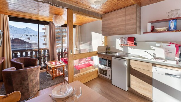Une cuisine chaleureuse avec des meubles en bois, un coin repas avec une table dressée pour quatre personnes, et un balcon offrant une vue sur des montagnes enneigées.