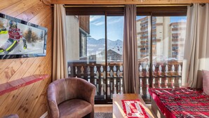 Une pièce chaleureuse avec un plafond en bois, un téléviseur diffusant un événement sportif, un fauteuil en cuir, une table en bois, un lit recouvert d’une couverture rouge et un balcon offrant une vue sur des montagnes enneigées.
