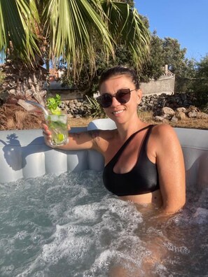 Una persona en un jacuzzi sosteniendo una bebida con lima, rodeada de follaje y un muro de piedra.