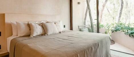 A bedroom with a large bed, an air conditioning unit, a view of trees through a window, and a bench outside.