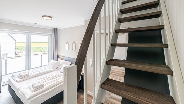A modern bedroom with a large bed, a wooden staircase, and a view of the outdoors. - Vacation home behind the dike for 6 pers. (Büsum)
