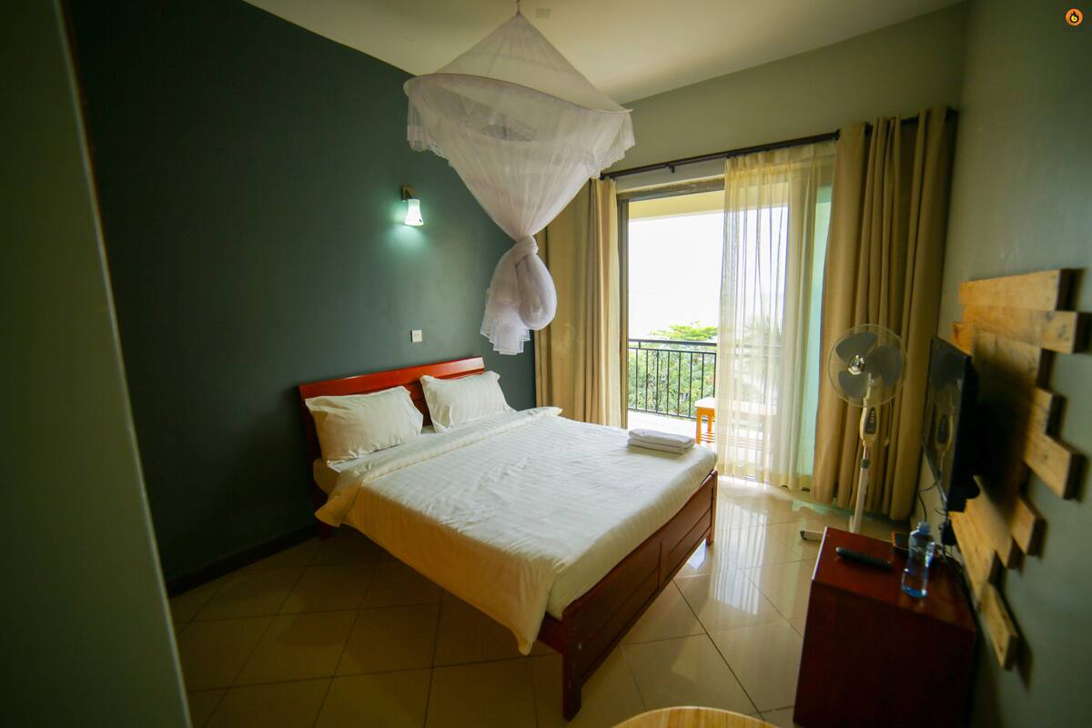 A hotel room with a bed, a bedside table with a remote control, a water bottle, and a ceiling fan.