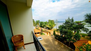 A balcony with a wicker chair, a view of a street with parked cars, and a body of water in the background. - Lakefront Hotel Entebbe (Entebbe)