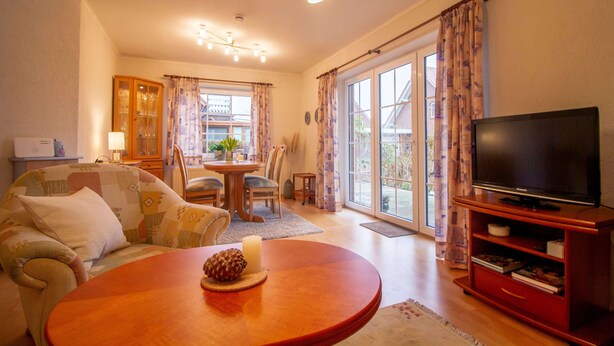 Een woonkamer met een bank, een houten salontafel, een televisie op een standaard en een eethoek met een tafel en stoelen.