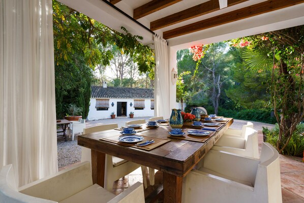 An outdoor dining area with a wooden table set for four, surrounded by white chairs, with a view of a white building and lush greenery in the background.