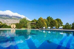 A clear blue pool with lounge chairs, surrounded by lush greenery and a mountain in the background.