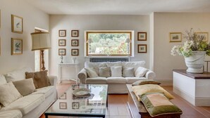 A living room with a beige sofa, a glass coffee table, and a white armchair. There are framed pictures on the walls and a window with a view of greenery. - 'Torre del Falco', a wonderful medieval tower in Spoleto (Spoleto)