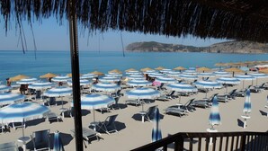 Une plage bordée de rangées de parasols et de chaises longues, une terrasse en bois et une vue sur la mer et les collines lointaines.