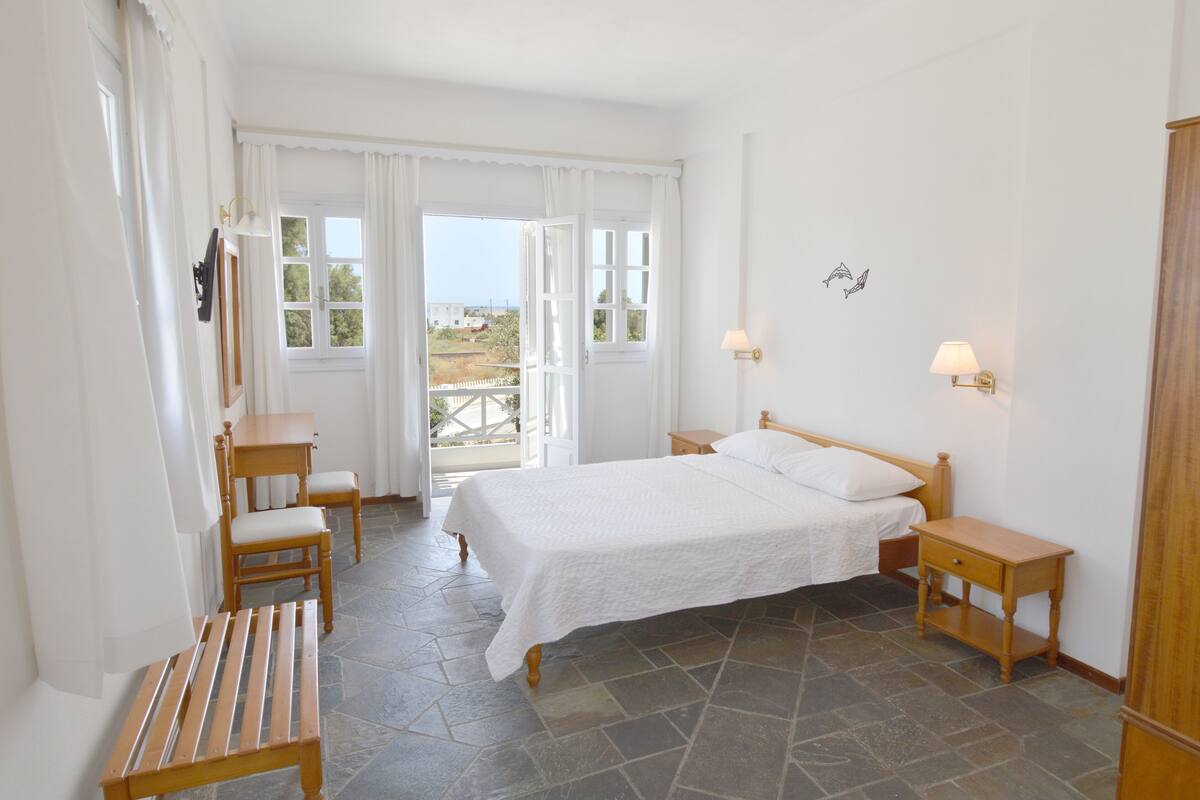 A bedroom with a bed, two bedside tables, a wooden bench, and a view of the outdoors through a large window.