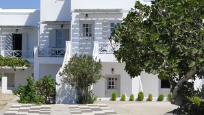 A white building with a checkered pavement, a tree, and neatly trimmed bushes. - Nostos Studio (Santorini)