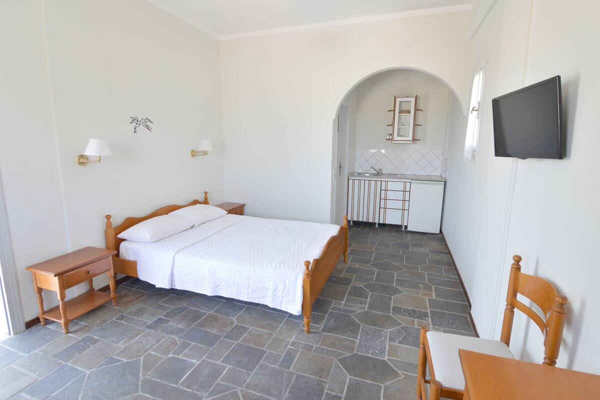 A bedroom with a bed, bedside tables, a chair, and a small desk with a chair. There is a TV mounted on the wall and a bathroom visible through an archway.
