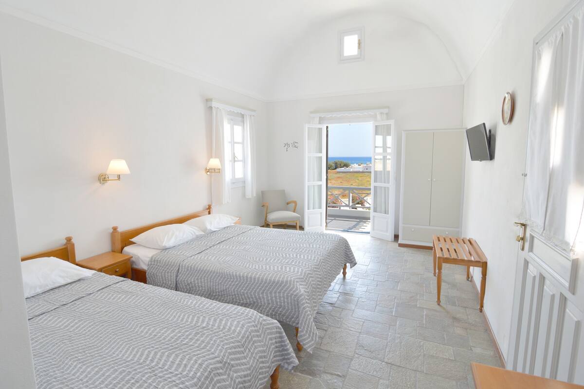 A hotel room with two beds, a wooden bench, a TV mounted on the wall, and a view of the outdoors through an open door.