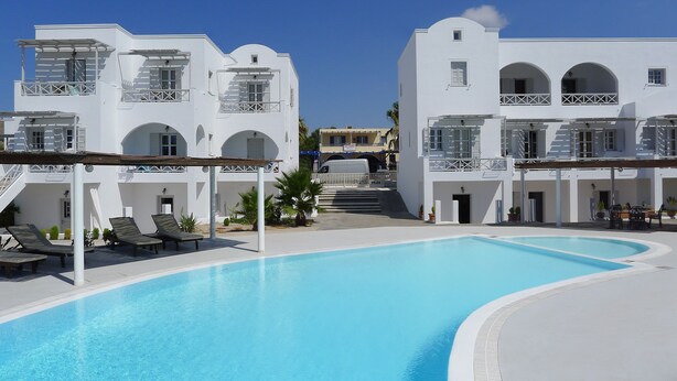A swimming pool area with white buildings, sun loungers, and a covered seating area. - Nostos Studio (Santorini)