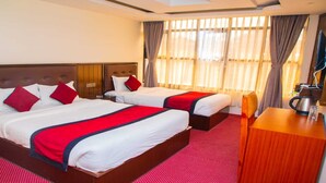 A hotel room with two beds, each with red and white bedding, a wooden headboard, a wooden desk with a kettle, and a large window with sheer curtains. - MeroStay004 (Kathmandu)