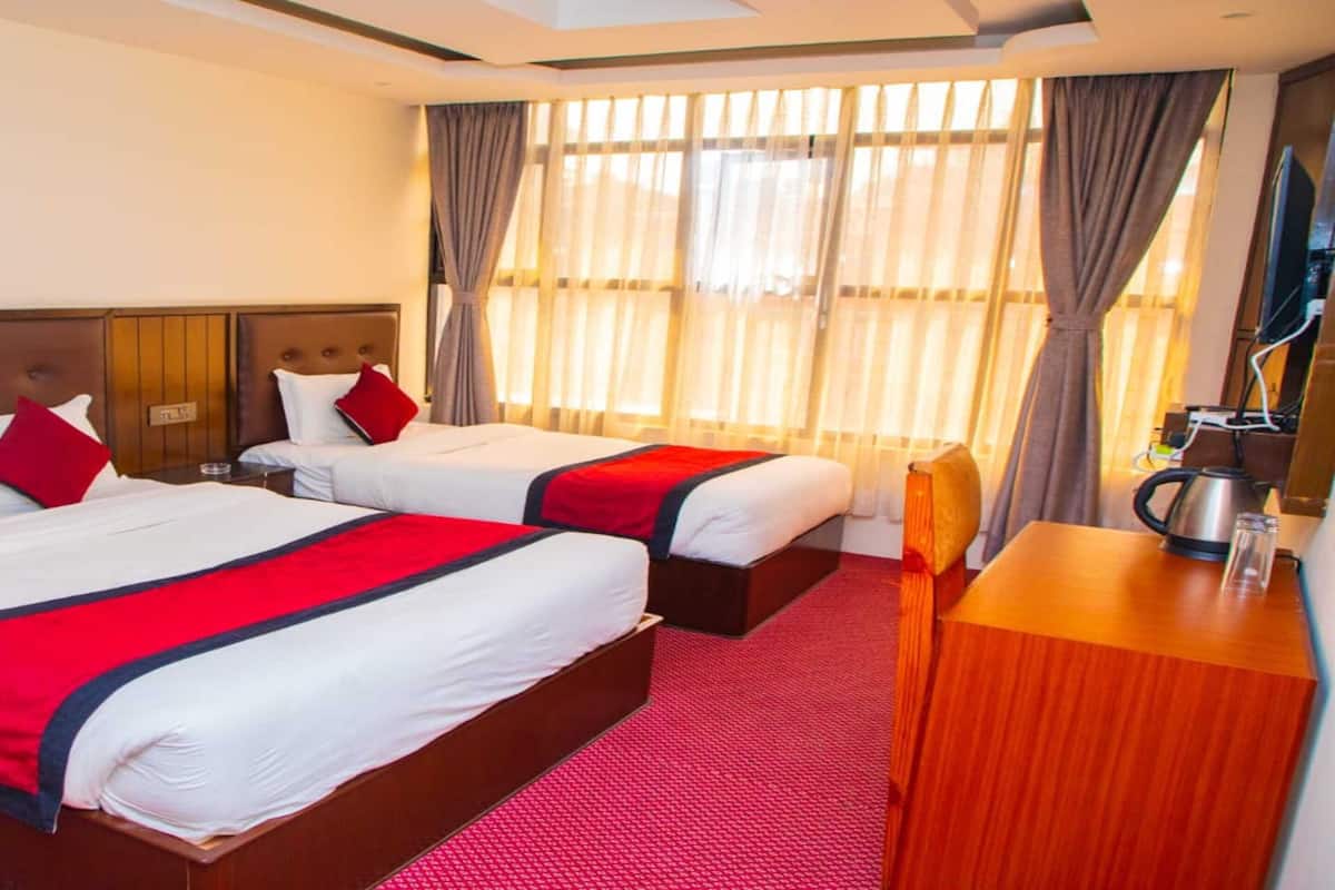 A hotel room with two beds, each with red and white bedding, a wooden headboard, a wooden desk with a kettle, and a large window with sheer curtains.