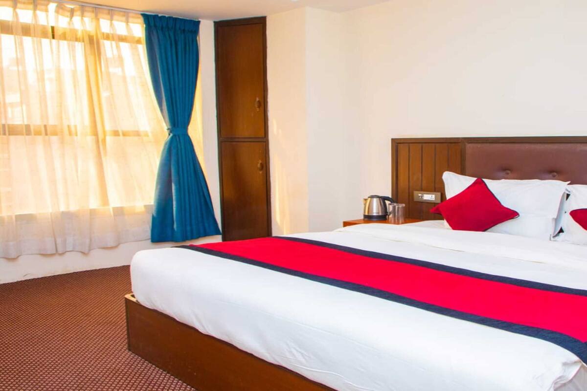 A hotel room with a large bed, a wooden headboard, a red and white bedspread, and a window with sheer curtains.
