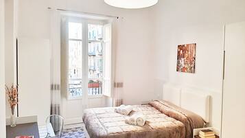 Um quarto com uma cama, uma mesa de cabeceira com livros, uma janela com vista para prédios e uma porta.