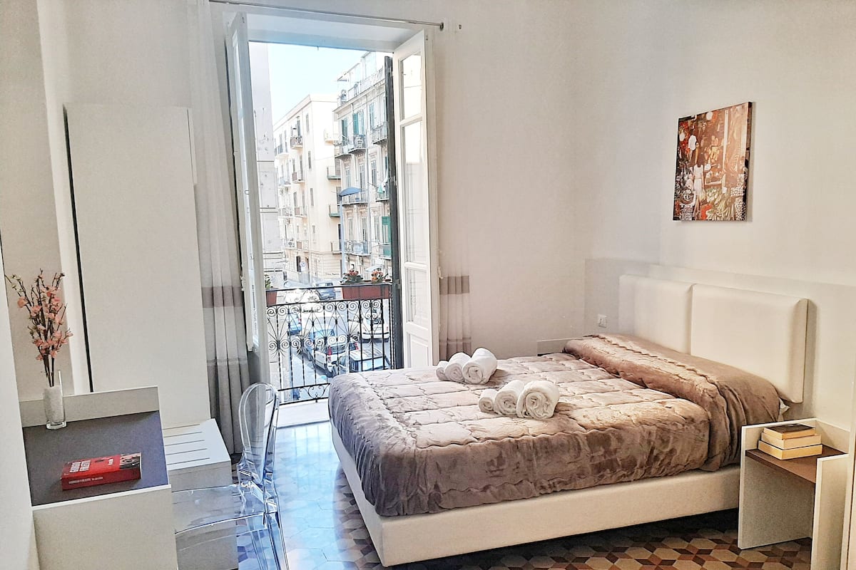 Une chambre à coucher avec un lit, une petite table de chevet, un miroir et une vue sur la rue, visible à travers une porte ouverte.