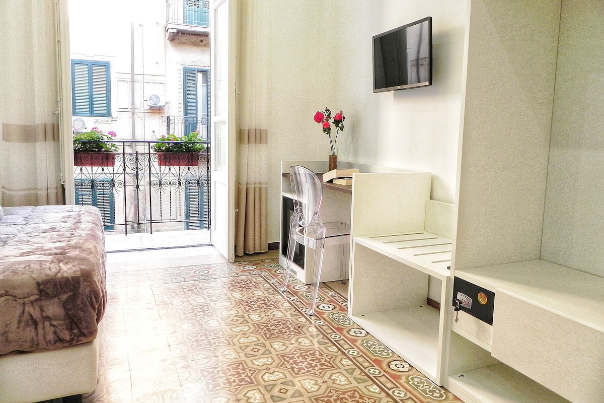 Une chambre avec un lit, un téléviseur fixé au mur, un radiateur et un balcon donnant sur des immeubles.
