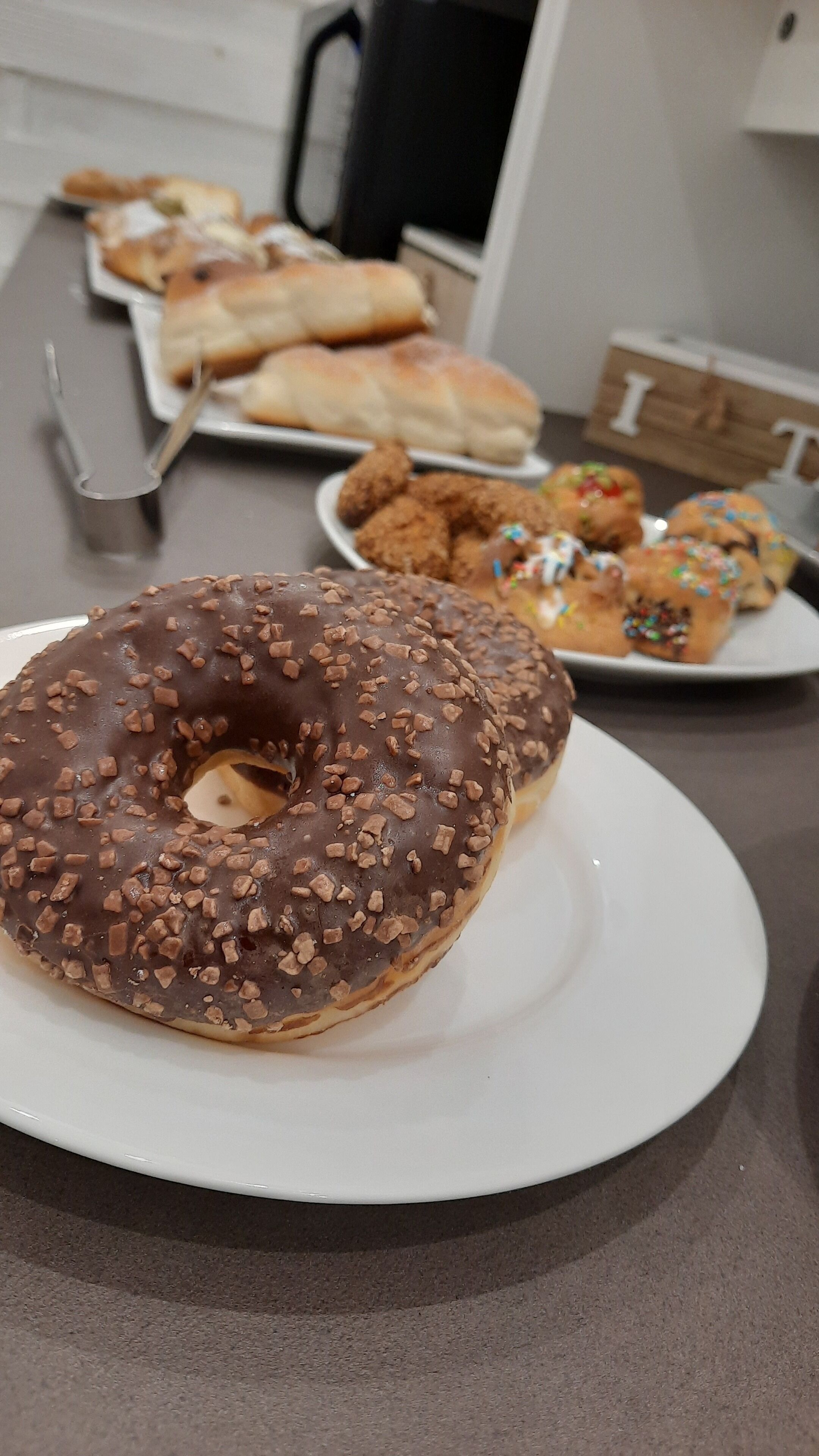 A variety of pastries on a table, including a chocolate-covered donut with nuts and a plate of fried chicken.