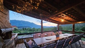 An outdoor dining area with a stone floor, wooden table, and chairs, overlooking a mountainous landscape.