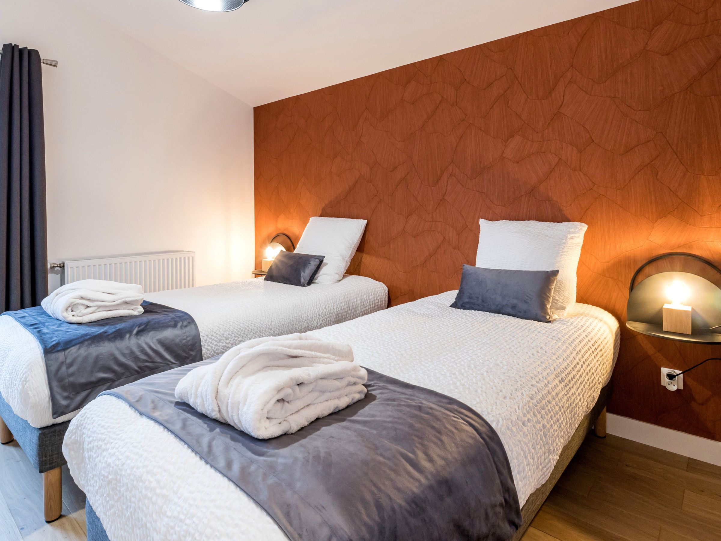 A hotel room with two beds, each with a white duvet, grey and white pillows, and a folded white towel. The room has a wooden headboard, a bedside table with a lamp, and a wall-mounted light fixture.