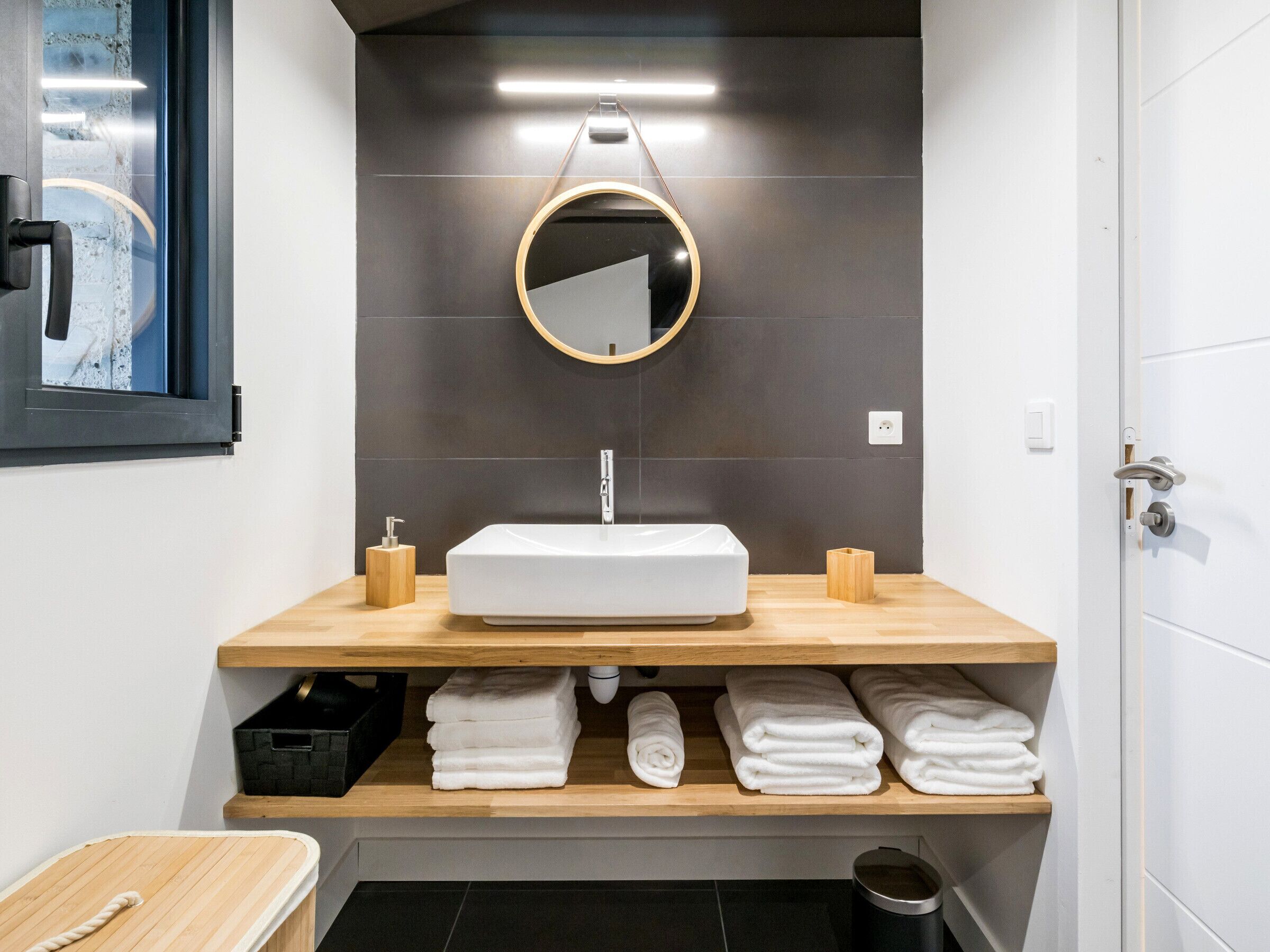 Un baño moderno con un lavamanos rectangular, un espejo redondo y un perchero con toallas dobladas.