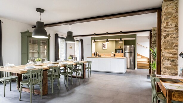 Un vaste espace de salle à manger avec une longue table en bois et des chaises vertes, une cuisine agrémentée d’un mur en pierre, et un escalier menant à un autre niveau.