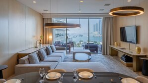 Een moderne hotelkamer met een groot raam met uitzicht op de stad, een zithoek met een gedekte tafel voor twee personen, en een lounge met banken en een televisie aan de muur.