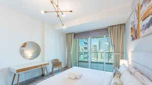 A modern hotel room with a large bed, a desk, a chair, and a view of the cityscape through the window. - Elegant 1 Bed Escape | Walk to Metro Station | JLT (Jumeirah Lake Towers)