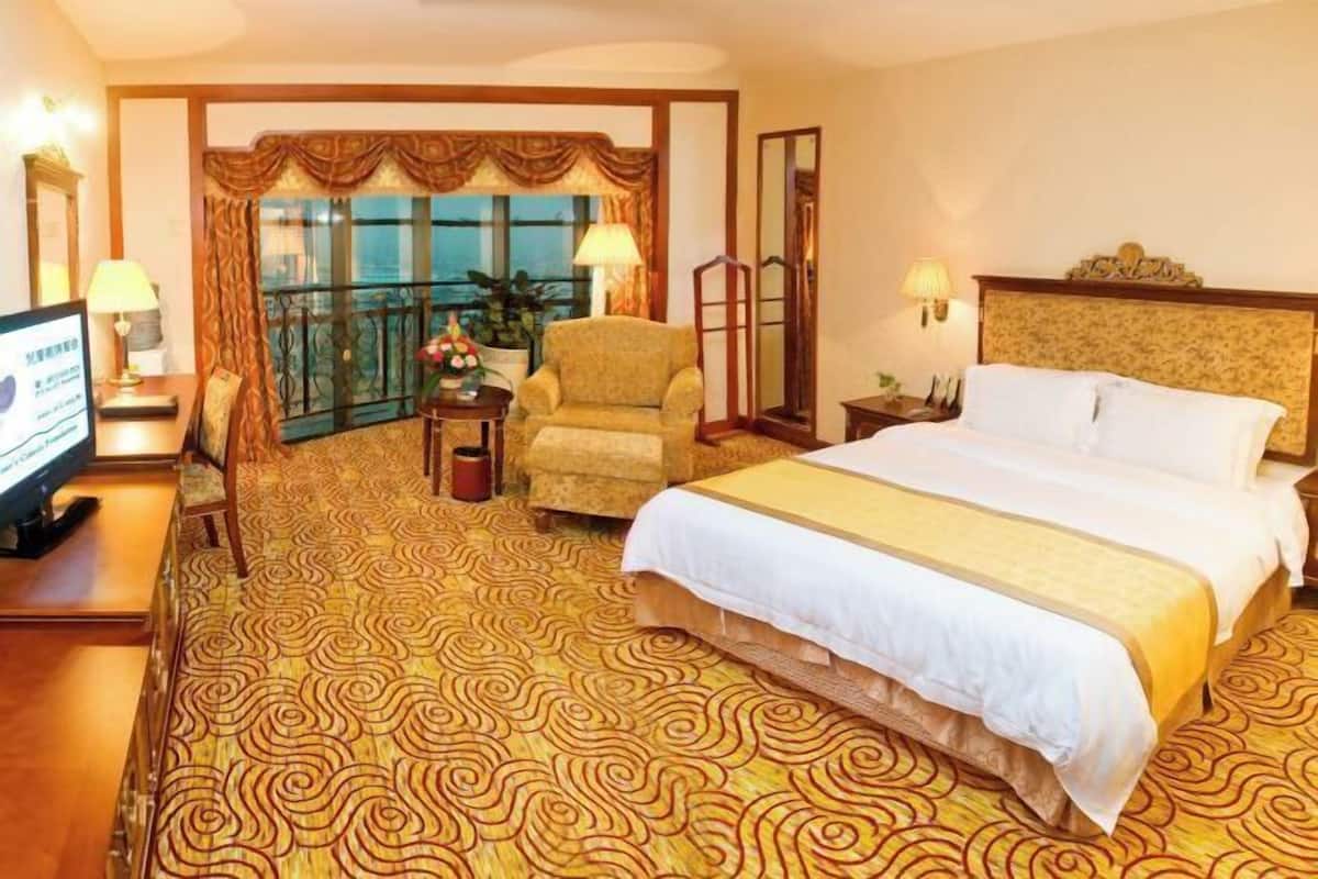 A hotel room with a large bed, a chair, a TV, a desk, and a balcony with curtains.
