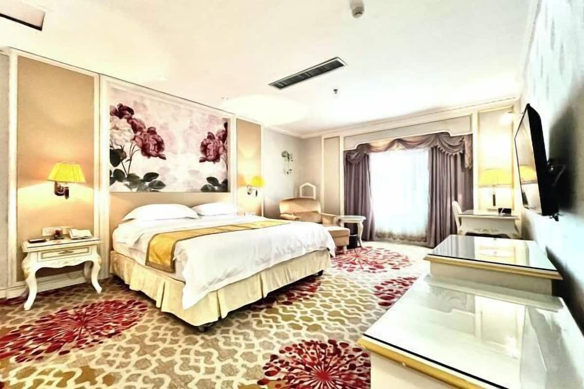A hotel room with a large bed, a desk, a chair, a TV, and a large floral painting on the wall.