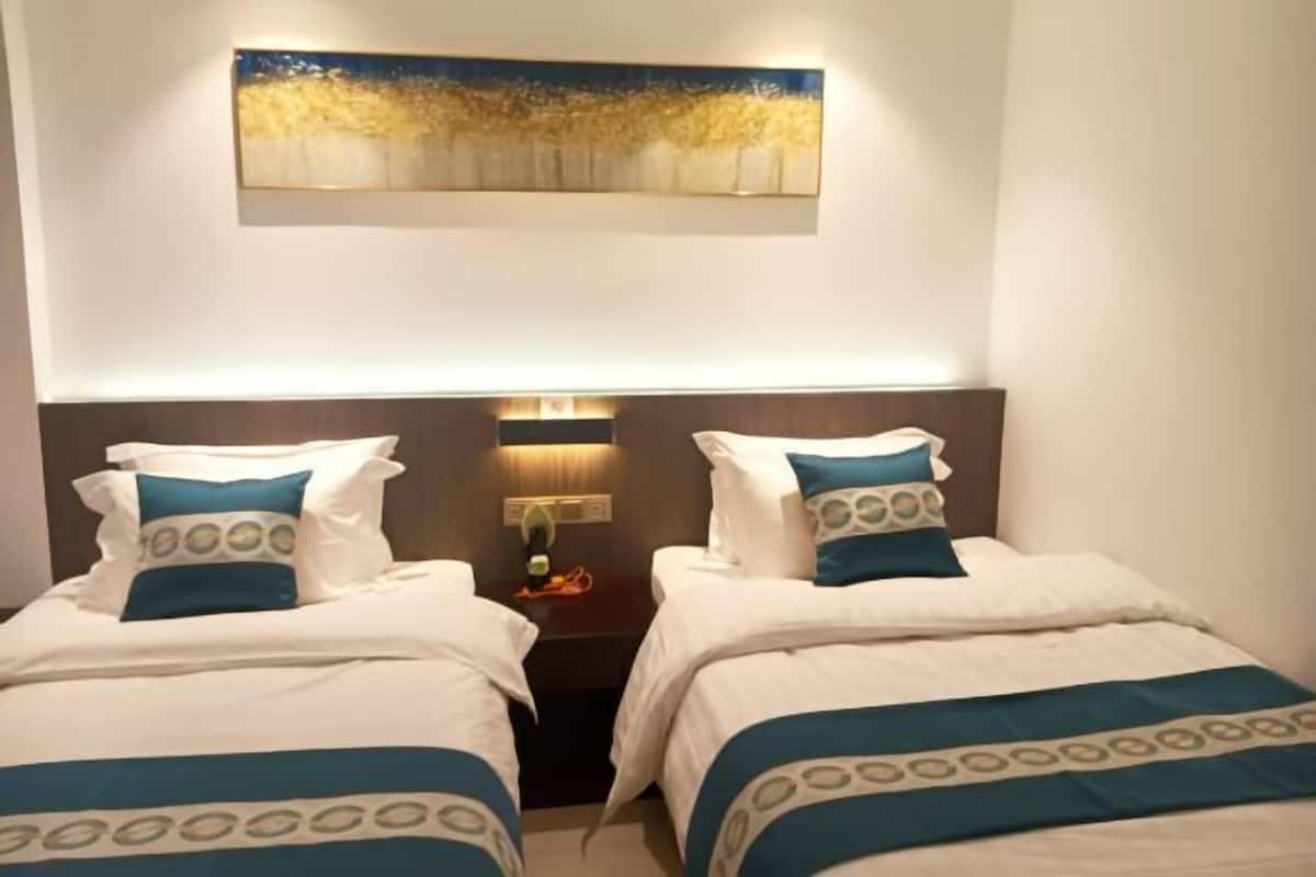 A hotel room with two beds, each with a blue and white bedspread, a nightstand with a lamp, and a painting above the beds.