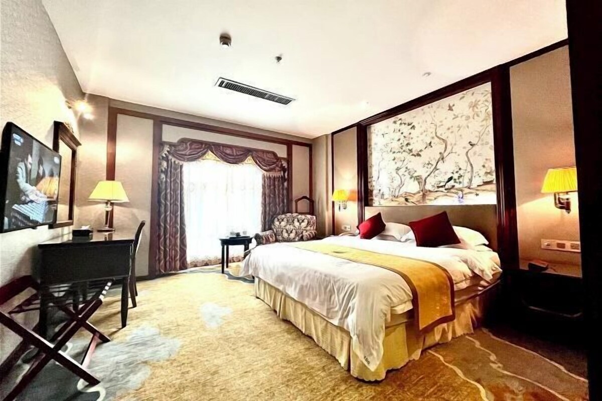 A hotel room with a large bed, two bedside tables with lamps, a desk, a chair, and a large window with curtains.