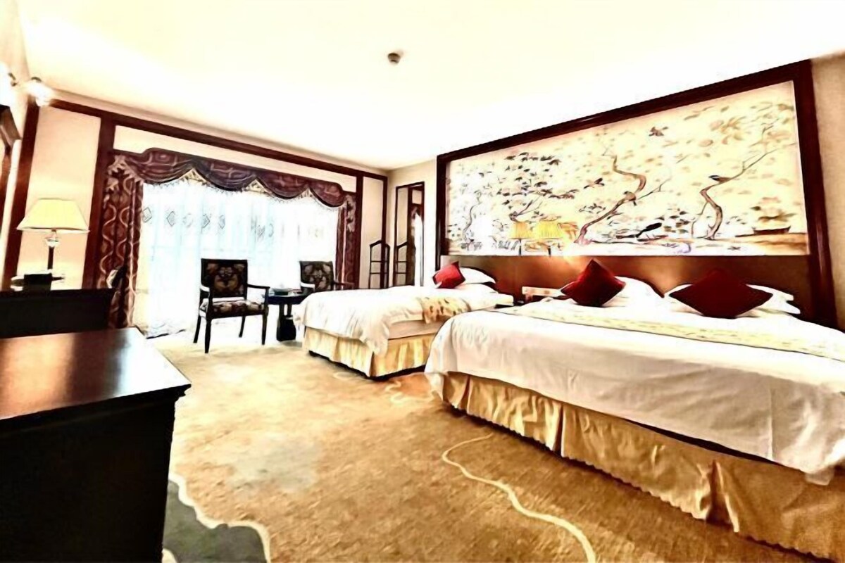 A hotel room with two beds, a desk, and a chair. There is a large painting on the wall and a window with curtains.
