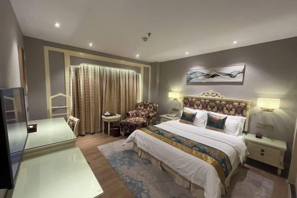 A hotel room with a bed, two bedside tables, a chair, a glass desk, and a large window with curtains.