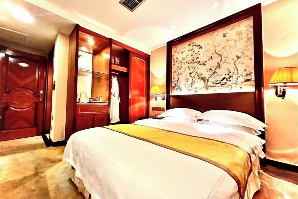 A hotel room with a large bed, wooden wardrobe, and a painting on the wall.
