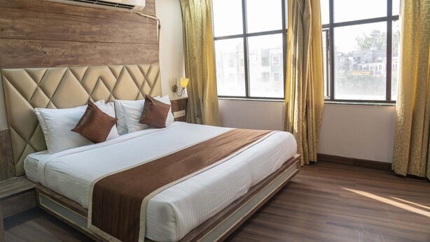 Quarto de hotel com uma cama grande, cabeceira de madeira e vista da cidade através de janelas amplas.