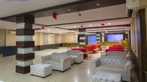 Um hall de hotel com sofás e pufes brancos, um balcão de recepção e uma área de refeições com mesas vermelhas e cadeiras amarelas.