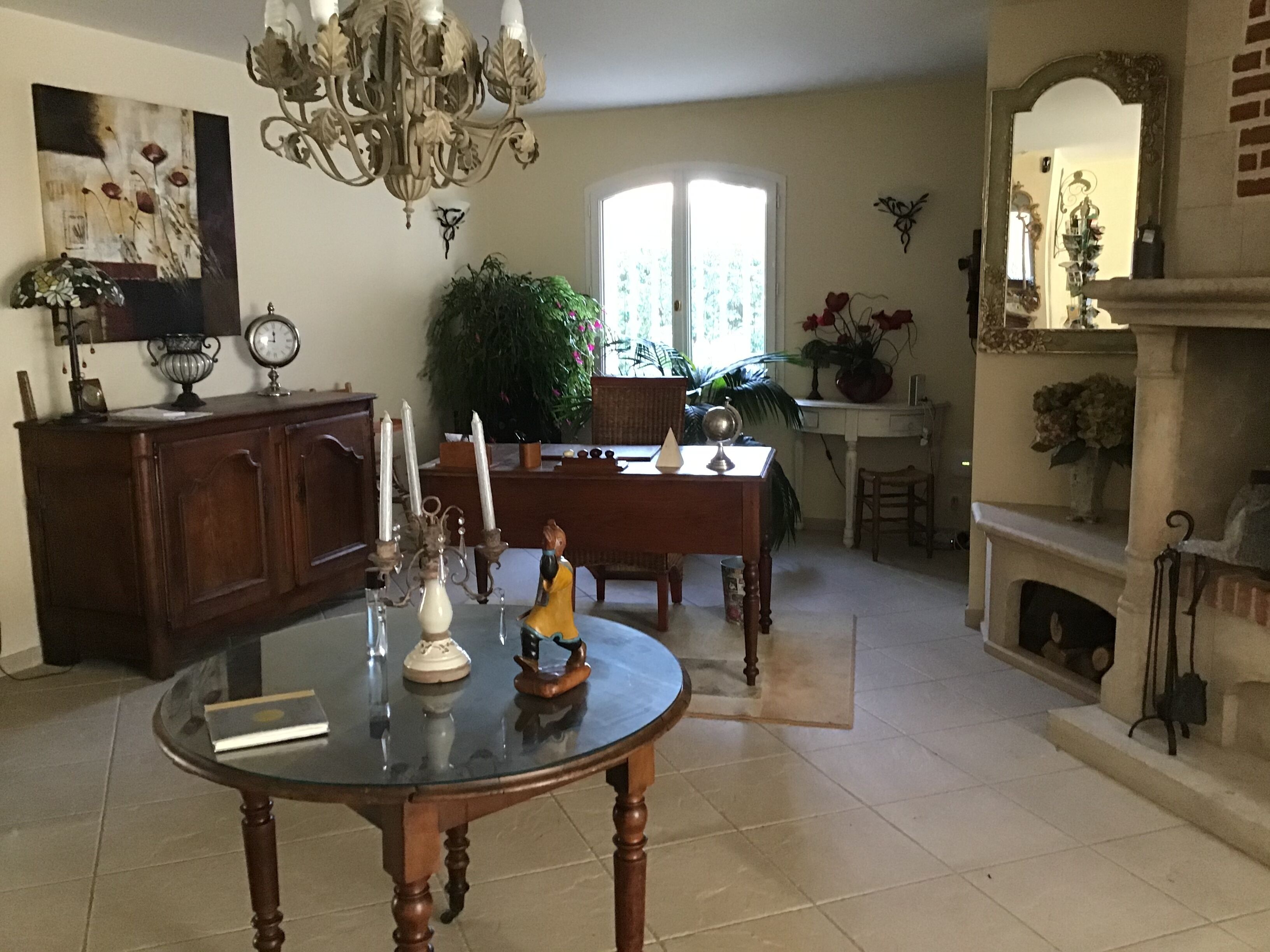 A dining area with a round table, chairs, a chandelier, a fireplace, and a large window.