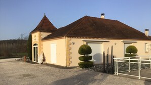 A two-story building with a brown tiled roof, a white garage door, and a small tower-like structure on the left. There are two neatly trimmed topiary bushes in front of the garage. - Pretty house with swimming pool in Périgord (Pazayac)