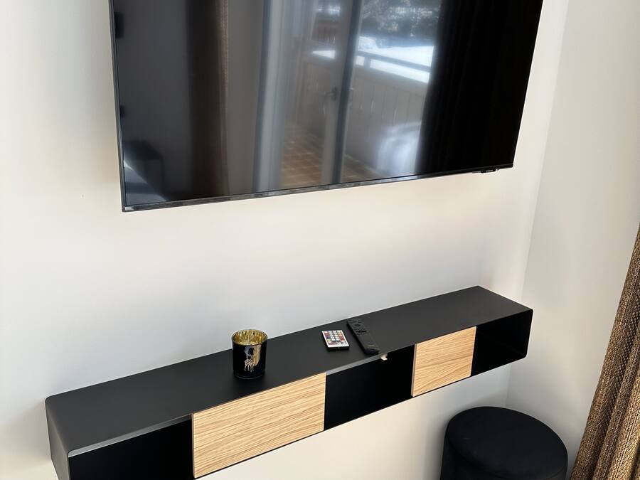 A modern living room with a flat-screen TV mounted on the wall, a black console table with a remote control, and a black ottoman.