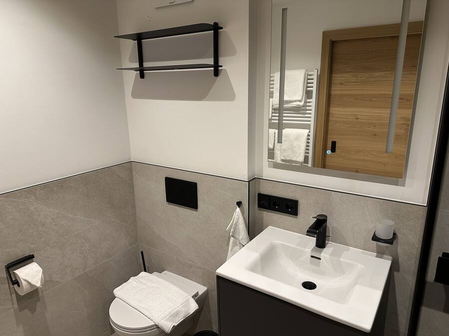 Un baño moderno con inodoro, lavamanos, toallero y un espejo que refleja una puerta de madera.