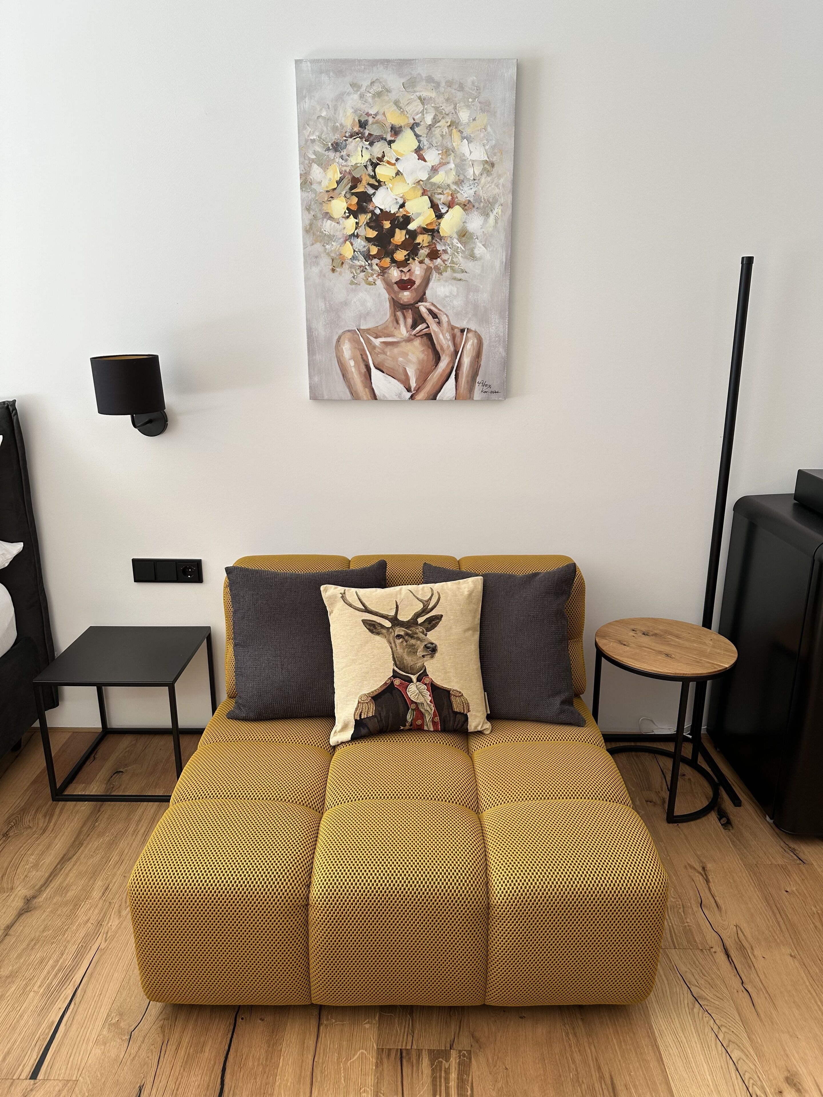 A modern living room with a yellow sofa, a deer pillow, a painting, and a black lamp.