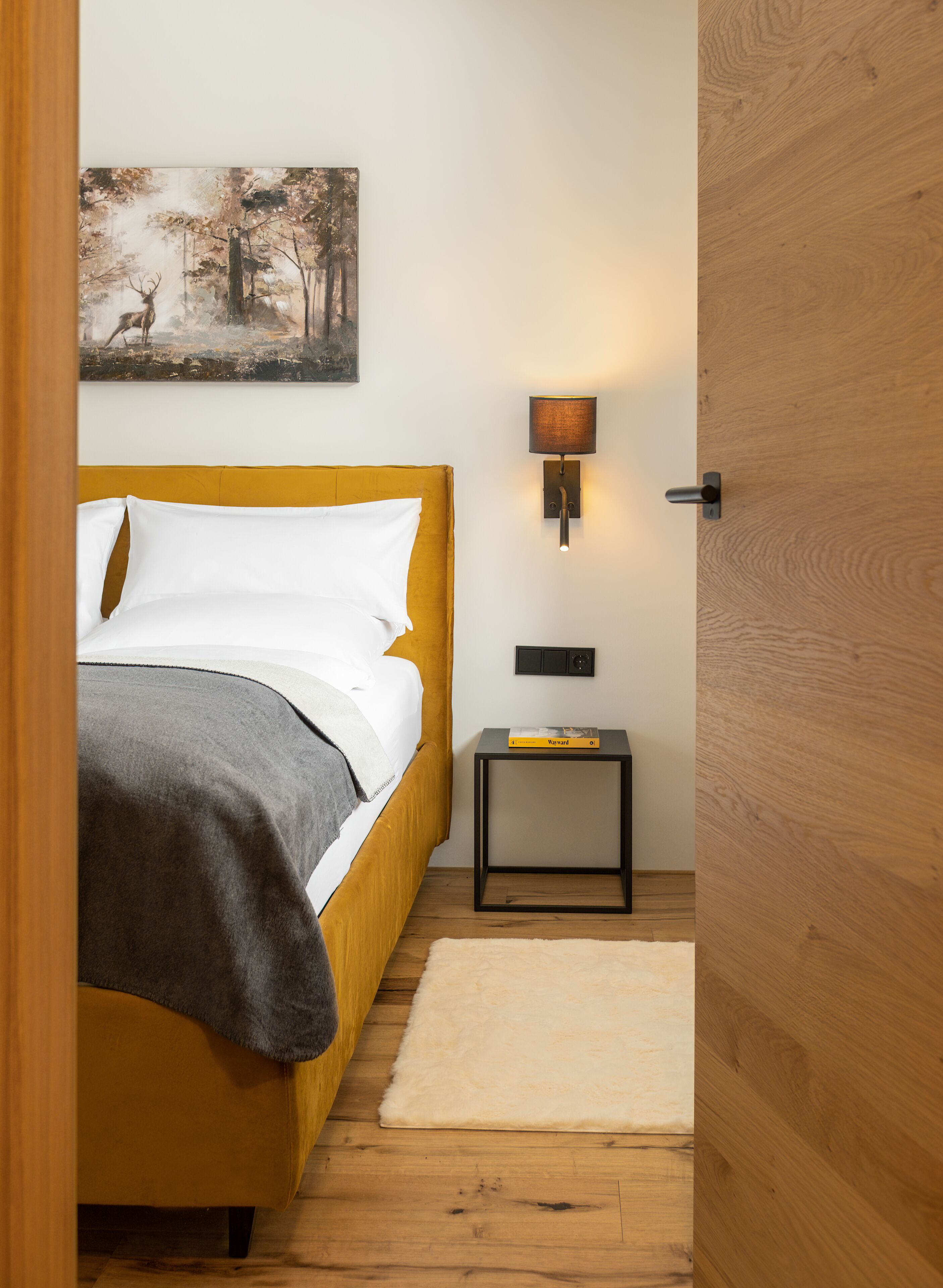 A bedroom with a bed, a nightstand, a lamp, a painting, and a wooden door.