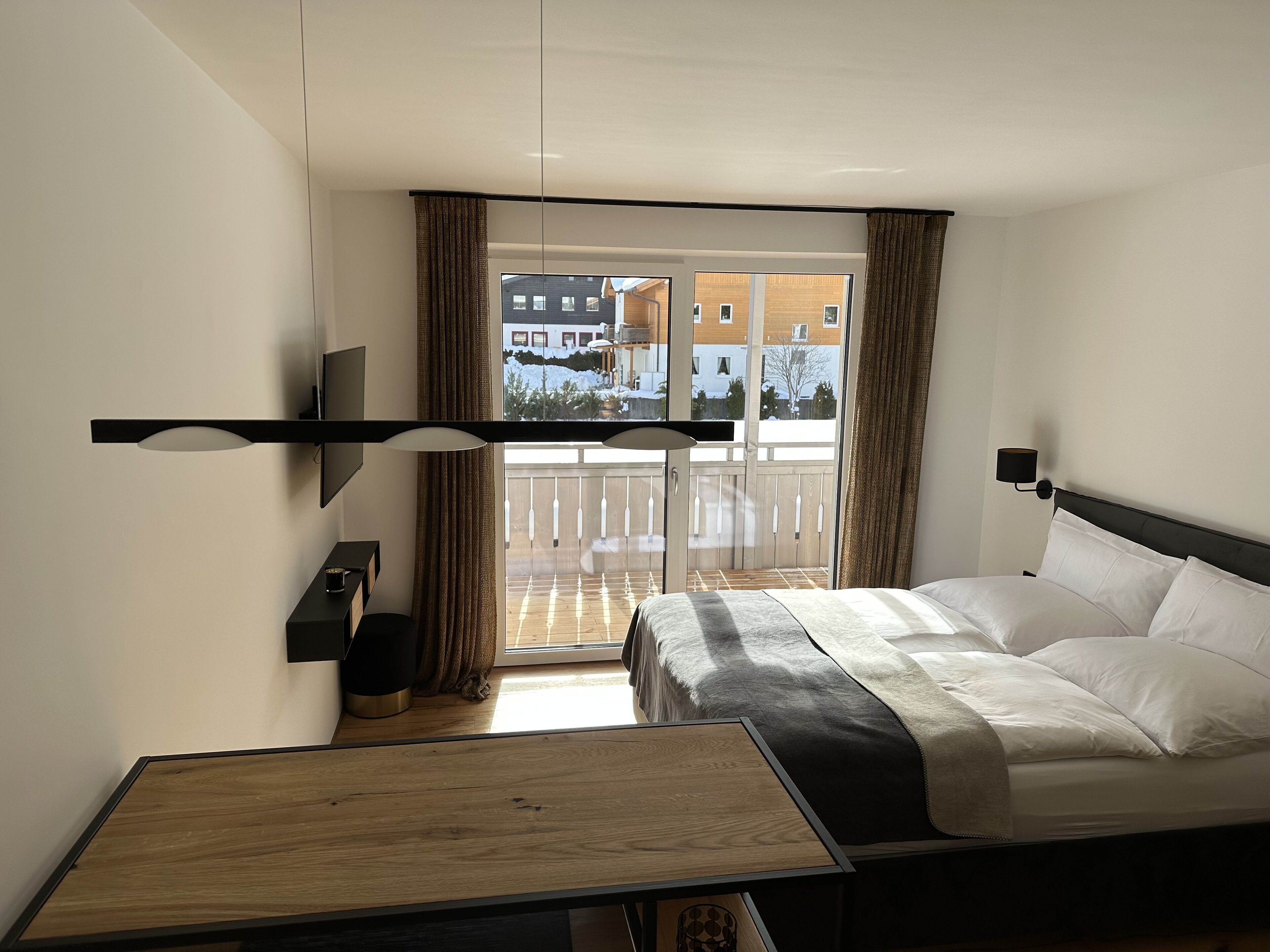 Habitación de hotel con cama, una mesita de madera, televisor y balcón con cortinas.